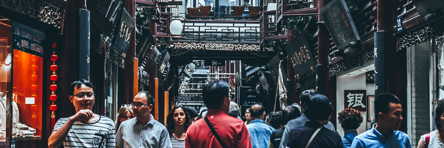 A bustling, narrow street market is shown, filled with people walking and browsing. Traditional Chinese architectural elements and shop signs are visible. The atmosphere is lively, with a mix of locals and tourists engaging in the vibrant marketplace environment.